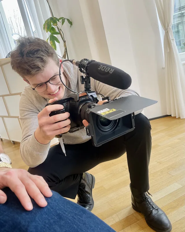 Eine Person mit Kamera und aufgesetztem Mikrofon bei einer Aufnahme im Innenraum.