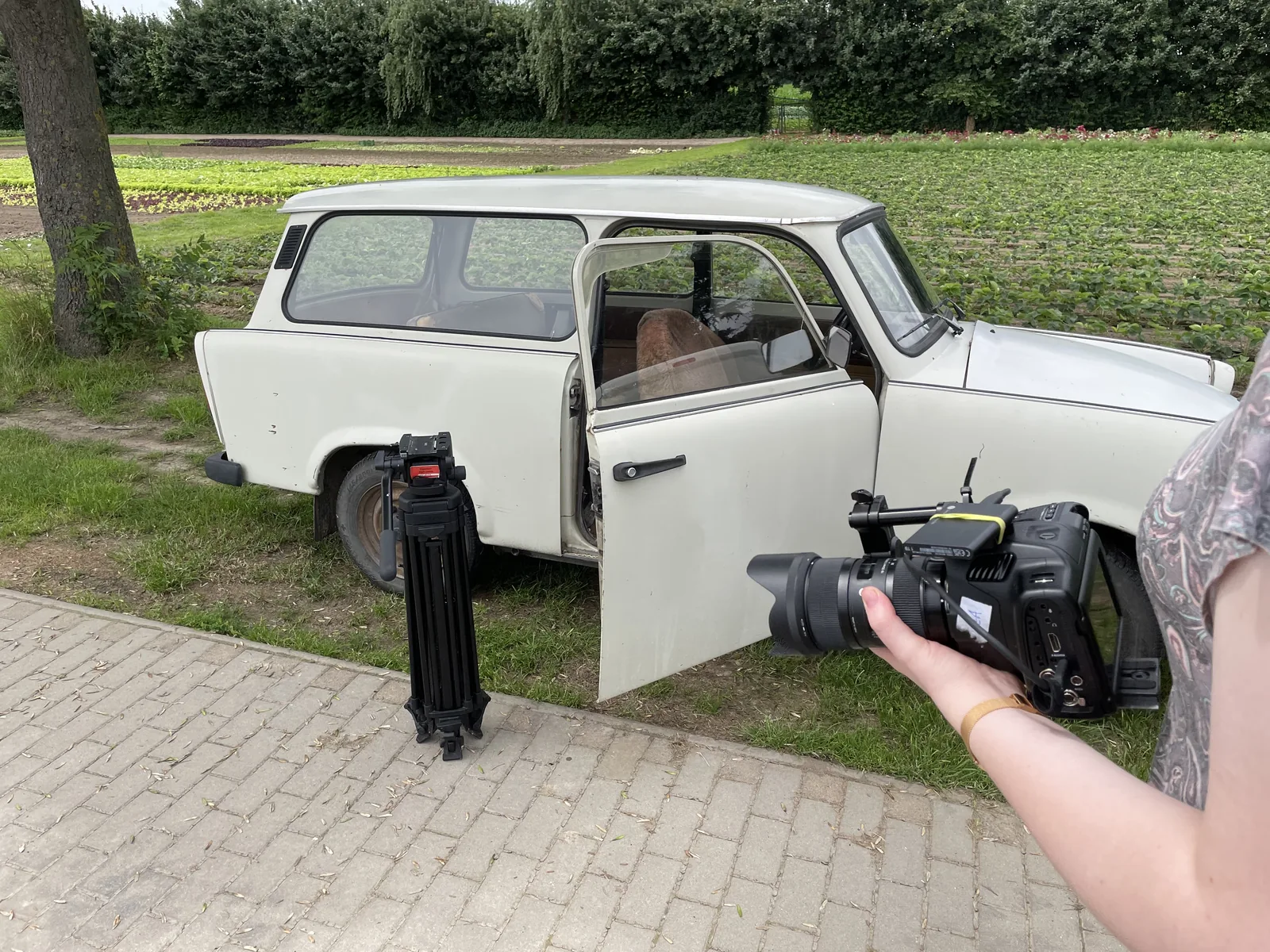 Kamera und Stativ bei einer Aufnahme neben einem weißen Fahrzeug auf dem Gartengelände.