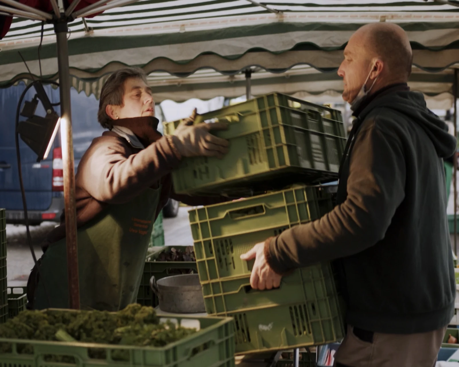 Zwei Menschen heben grüne Transportkisten an einem Marktstand.