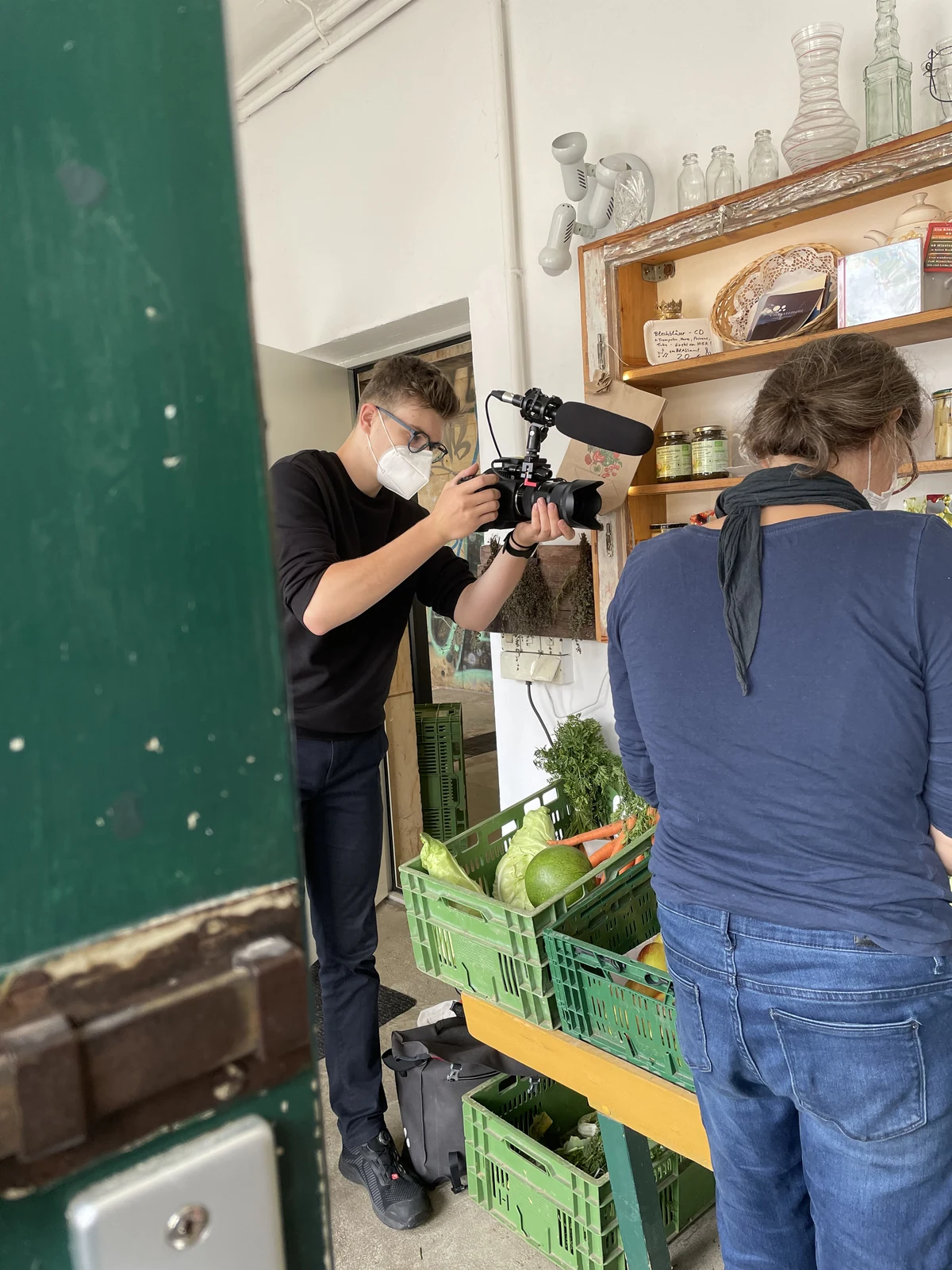 Ein Kameramann filmt zwischen Türrahmen und Gemüsekisten im Laden.