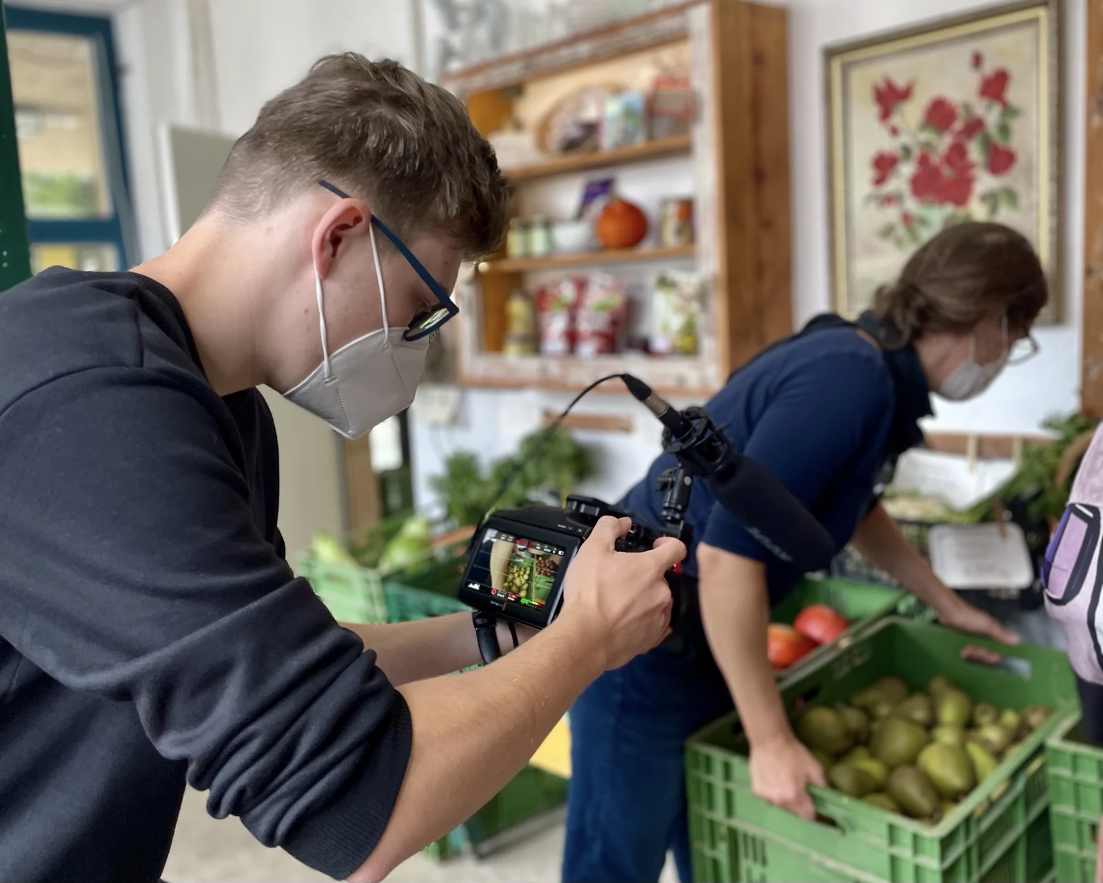 Ein Kameramann nimmt eine Person beim Arbeiten an Gemüsekisten im Laden auf.