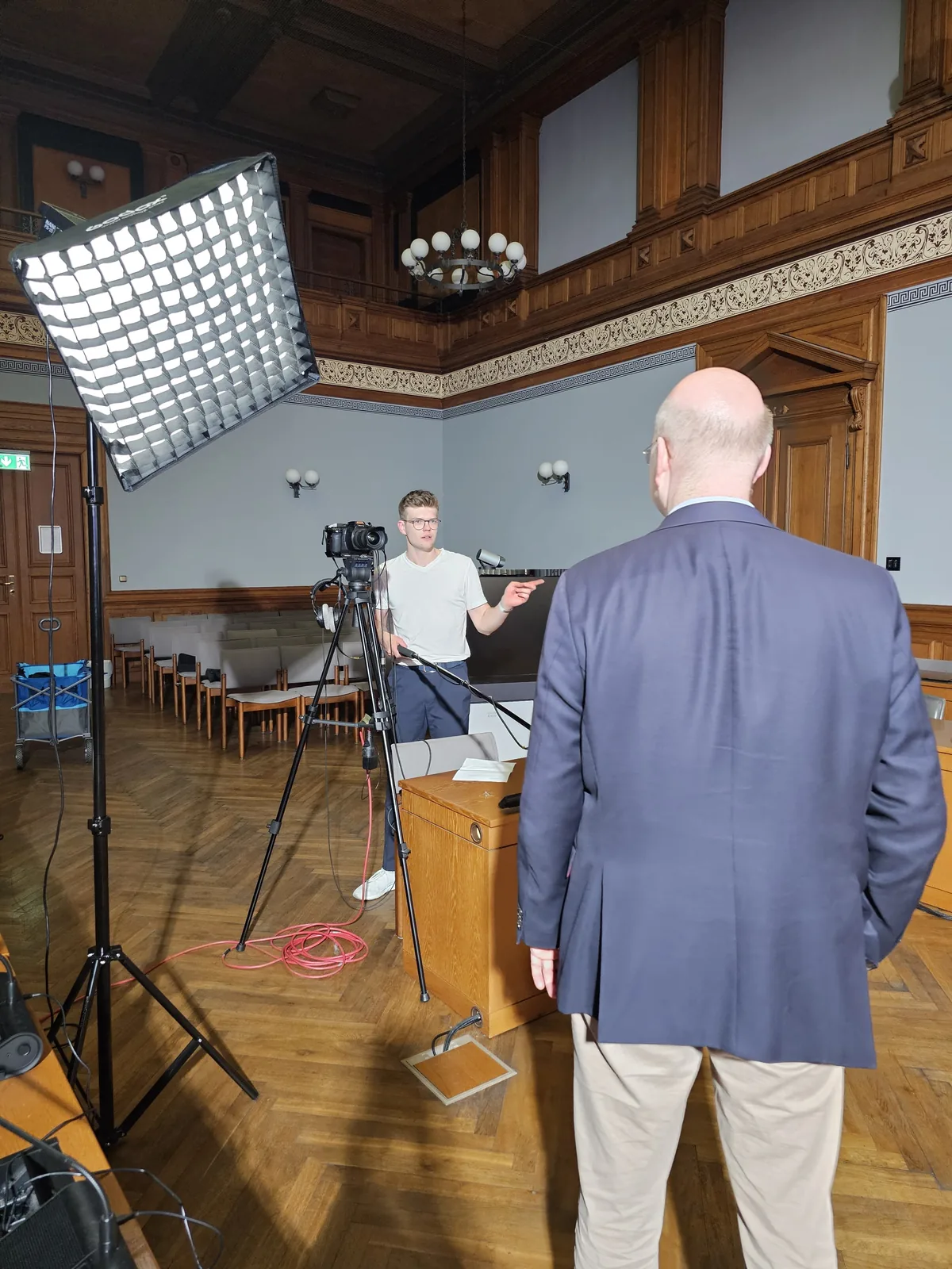 Blick über eine Gesprächsperson auf Kamera und Beleuchtung in einem holzvertäfelten Saal.